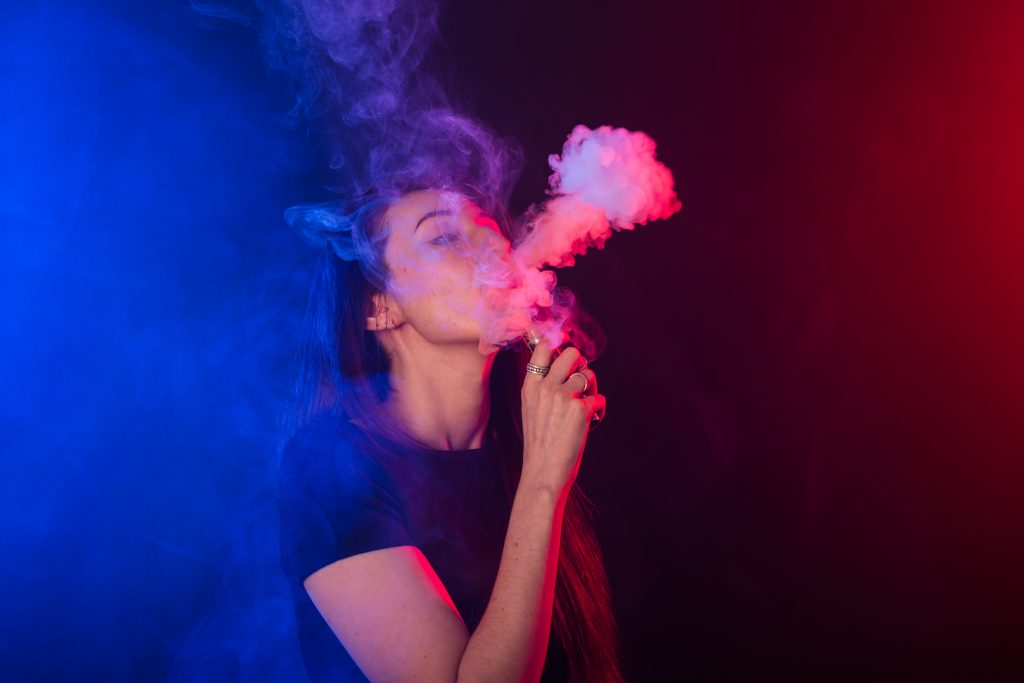 A woman exhales colorful vapor in a dimly lit space, with blue and red lights creating an atmospheric backdrop.
